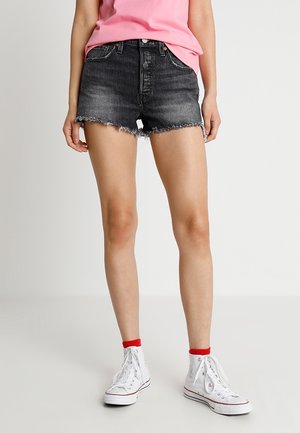 Shorts en jean délavés noirs avec un ourlet effiloché, associés à des baskets montantes blanches et des chaussettes accentuées de rouge, portant un haut décontracté rose.