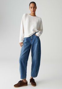 Donna in piedi con mano in tasca che indossa un maglione bianco ampio, jeans larghi di denim blu e sneakers marroni, su sfondo semplice.