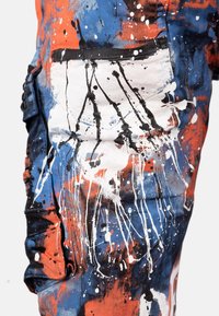 Pantalons cargo en denim éclaboussés de peinture, présentant un mélange de teintes bleues, orange et blanches. Détails de poches texturés notables avec des accents noirs.