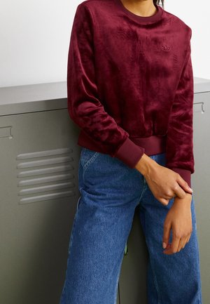 Osoba ubrana w bordową, welurową bluzę z długim rękawem z logo oraz niebieskie, wysokowalne jeansy, stojąca obok szarej szafki.