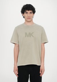 Beige bomull t-skjorte med korte ermer, med en lett rundet halslinje og en stor, dempet grønn "MK" logo på forsiden.