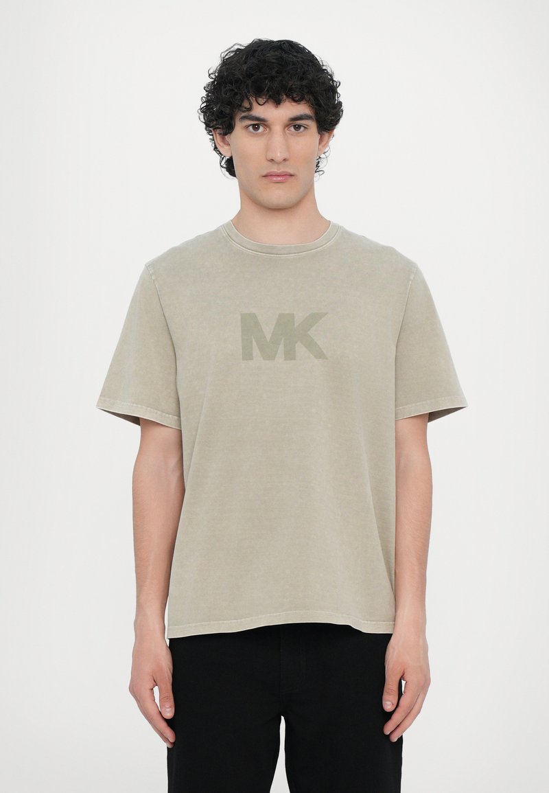 Beige bomull t-skjorte med korte ermer, med en lett rundet halslinje og en stor, dempet grønn "MK" logo på forsiden.