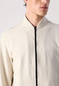 Uomo che indossa una giacca a zip color crema con texture e colletto a coste, rivolto leggermente a destra, mostrando il torso superiore e la parte bassa del viso.