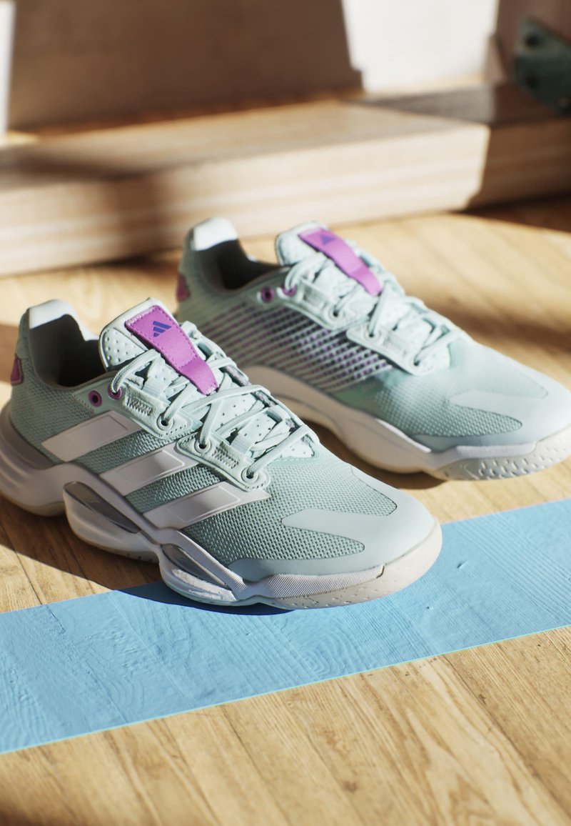 Zapatillas deportivas de color verde claro con acentos blancos, parte superior de malla texturizada, detalles y cordones morados, y suelas acolchadas, posicionadas sobre una superficie de madera.