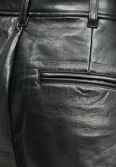 Detailný záber na čierne kožené nohavice, zobrazujúci textúrovaný materiál, bočný šev, pás s pútkami na opasok a horizontálny otvorený vrecko.