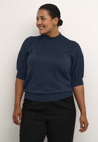 Maglione corto blu navy a maniche corte con collo alto a coste, tessuto texturizzato e maniche a sbuffo, abbinato a pantaloni neri.