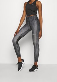 Femme portant un haut noir sans manches, un legging Nike noir et gris à motifs, et des chaussures de course Nike noires, debout devant un fond uni.
