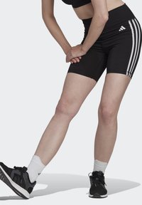 Czarne spodenki sportowe z trzema białymi poziomymi paskami po bokach, zestawione z czarnymi trampkami i białymi skarpetkami. Gładka tekstura materiału.