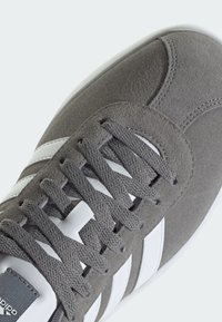 Zapatilla de ante gris con detalles y cordones de cuero blanco; presenta un diseño clásico de tres franjas y una superficie texturizada. Estilo simple y de perfil bajo.