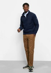 Marineblauer Fleecepullover mit halbem Reißverschluss, Brusttasche und geripptem Saum. Kombiniert mit sandfarbenen Chinos und dunkelgrünen Sneakern.