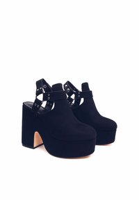 Scarpe platform in suede nero con tacco alto. Presentano un design con cinturino e dettagli metallici, e un tallone aperto. Texture liscia in tutto il corpo.