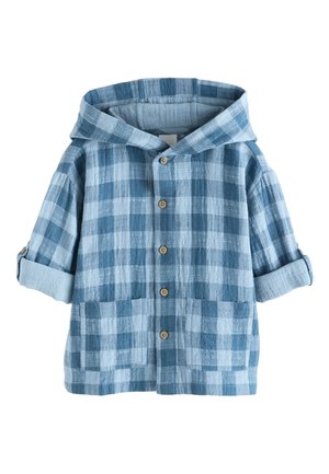 Barn med krøllet hår iført en blå ternet jakke, hvid skjorte, beige bukser og hvide sneakers, står mod en ensfarvet beige baggrund.