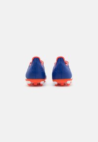 Botas de fútbol azules con detalles en naranja, con una parte superior sintética suave, cuello acolchado y suela blanca con tacos cónicos.