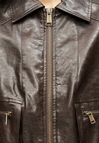 Καφέ δερμάτινο μπουφάν με ψηλό γιακά, εμπρόσθιο φερμουάρ και δύο τσέπες με φερμουάρ, διακοσμημένες με χρυσές λεπτομέρειες.