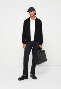 Zwarte gebreide bomberjack met textuurpatroon, witte polo, marineblauwe broek, zwarte schoenen en een gestructureerde zwarte schoudertas.
