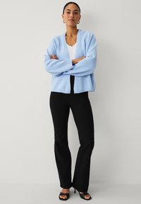 Cardigan tricoté bleu clair avec une coupe décontractée, poignets côtelés et devant ouvert, associé à un pantalon noir évasé et des chaussures noires à fines lanières.