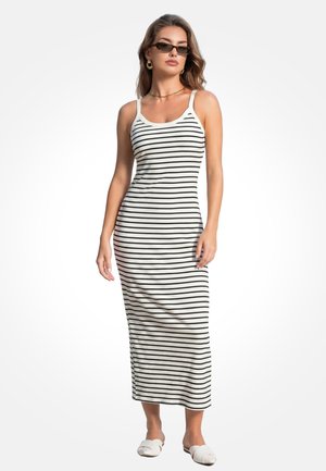 Donna che indossa un maxi abito senza maniche a righe bianche e nere, occhiali da sole, orecchini a cerchio dorati, collana e scarpe bianche slip-on, in piedi su sfondo bianco.