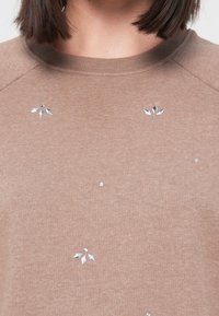 Gros plan sur une personne portant un pull beige avec de petites incrustations de gemmes argentées disposées en motifs floraux près de l'encolure et de la poitrine.