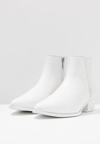 Botines blancos de tobillo hechos de cuero suave con punta puntiaguda, cremallera lateral y un pequeño tacón bloque. Diseño minimalista, sin patrones.