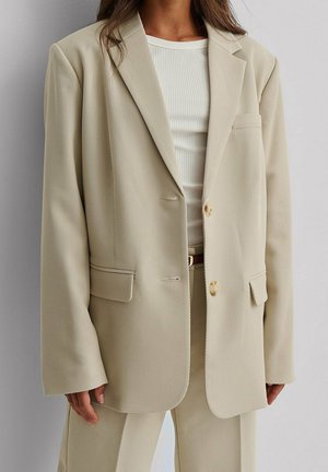 Kobieta w beżowej, dopasowanej marynarce i pasujących spodniach, nosząca biały prążkowany top na jednolitym tle.