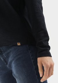 Zwarte langemouwen shirt met een textuurafwerking, inclusief een zoomlabel en een paar donkere jeans, met focus op de detail van de mouw en de pasvorm.