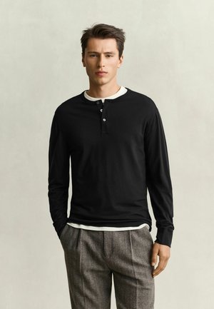 Giovane uomo in piedi con le mani in tasca che indossa una camicia henley nera a maniche lunghe sopra una maglietta bianca e pantaloni grigi.