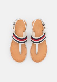 Un paio di sandali color sabbia con fasce per le dita bianche e bande in tessuto a righe rosse, blu navy e bianche, caratterizzati da piccole fibbie su uno sfondo bianco.