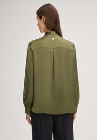 Blusa in satin verde oliva con collo alto, maniche lunghe e una piega centrale sul retro. Presenta un bottone decorativo nella zona del collo.