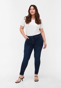 Jeans oscuros ajustados de talle alto, combinados con una camiseta blanca ajustada. La modelo lleva tacones negros de tiras. Tejido suave, con un detalle mínimo.