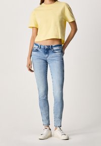 Camiseta de algodón amarilla recortada combinada con jeans skinny en un tono azul claro y zapatillas blancas. Los jeans tienen un desvanecimiento sutil y un diseño clásico de 5 bolsillos.