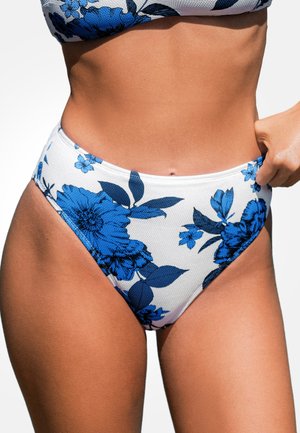 Kobieta w białych majtkach od bikini z wysokim stanem z dużym niebieskim kwiatowym wzorem, regulująca boczny pasek prawą ręką.