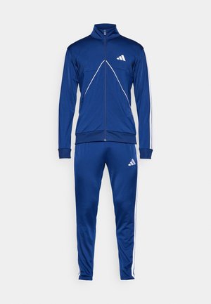 Tuta blu realizzata in un tessuto liscio e elasticizzato. Giacca con zip con accenti bianchi e logo, abbinata a pantaloni con vita elastica e strisce.