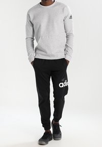 Sweatshirt gris à manches longues avec col rond et logo Adidas noir, associé à un jogging noir avec un logo blanc, le tout en tissu doux.