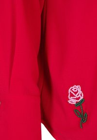 Sarkana blouse ar teksturētu audumu, kurā piedāvāta rožu izšūšana ar pērlītēm uz piedurknes rozā, baltā un zaļā krāsā. Vienkārša apaļa kakla izgriezuma forma.