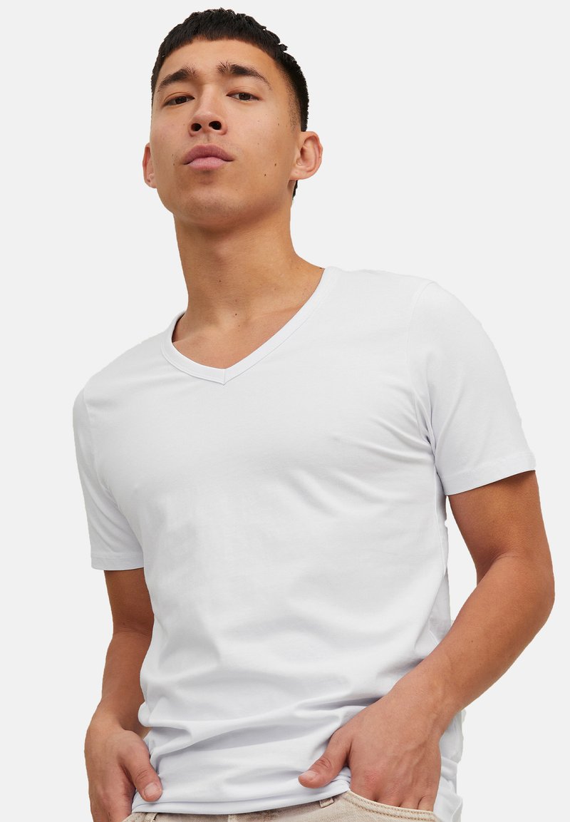 Jeune homme portant un t-shirt blanc uni à col en V, debout les mains dans les poches, regard confiant sur un fond neutre.