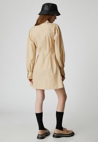 Hellbeiges Kleid mit puffartigen Ärmeln, geknöpften Manschetten und einem ausgestellten Rock. Kombiniert mit einem schwarzen Bucket-Hut und Schuhen mit einer groben Sohle.