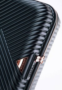 Valise rigide noire à texture côtelée, avec une fermeture éclair et des cadenas en rose doré, ornée d'une plaque logo triangulaire WITTCHEN.