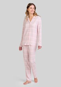 Mujer de pie descalza, vistiendo una camisa de pijama de manga larga a cuadros rosa claro y pantalones a juego, frente a un fondo liso.