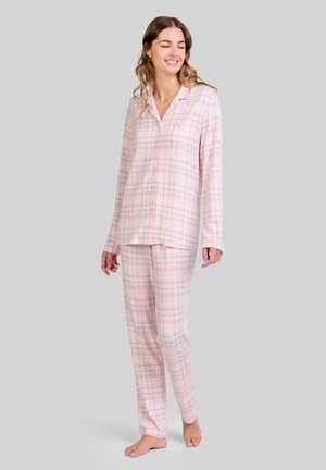 Mujer de pie descalza, vistiendo una camisa de pijama de manga larga a cuadros rosa claro y pantalones a juego, frente a un fondo liso.
