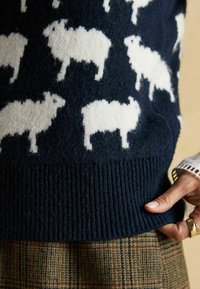 Tumedast villane sviiter, millel on valged lambamustrid, soonikäär ja vabalt langev lõige, koos ühelt poolt nähtava ruudulise seeliku alumise servaga.