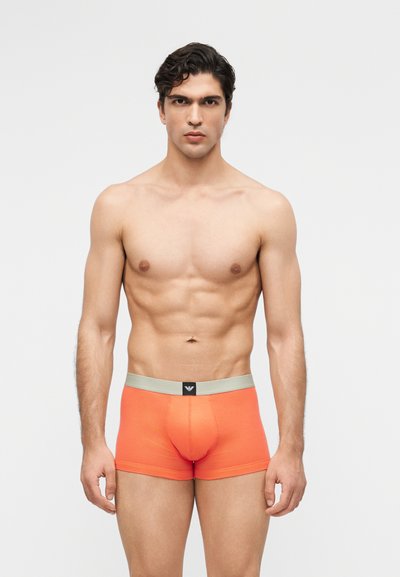 Muskelöser Mann mit dunklem Haar, der leuchtend orangefarbene Boxer-Shorts mit grauem Bund trägt und vor einfarbigem Hintergrund direkt zur Kamera steht.