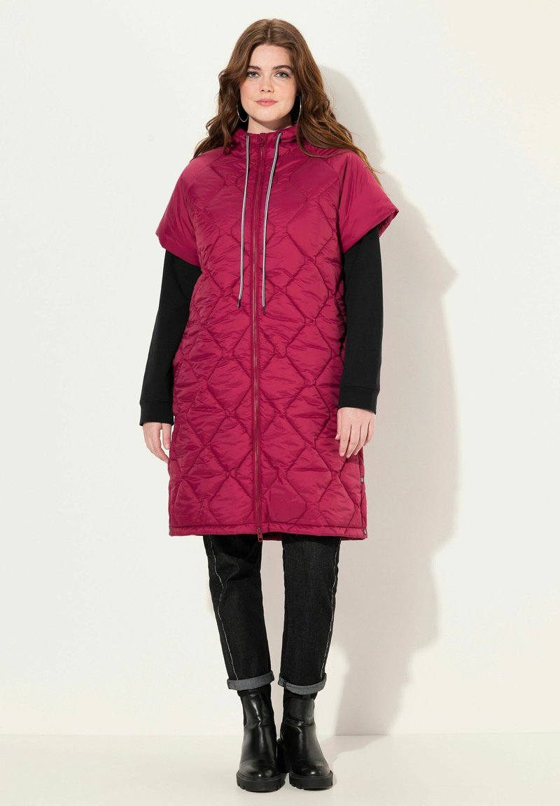 Překládaná, bezrukávová, burgundská vesta s vysokým límcem a zapínáním na zip. Nositá přes černé dlouhé rukávy, v kombinaci s tmavými džíny a černými botami.