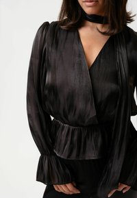 Melna satīna blūze ar apvēršanu, garām plandošām piedurknēm un pepluma jostasvietu. Iekļauts auduma elements ar sasienamu detaļu apkaklē.