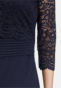 Robe bleu marine foncé avec corsage et manches en dentelle florale, manches en dentelle trois-quarts laissant apparaître la peau en dessous, et détail plissé à la taille.