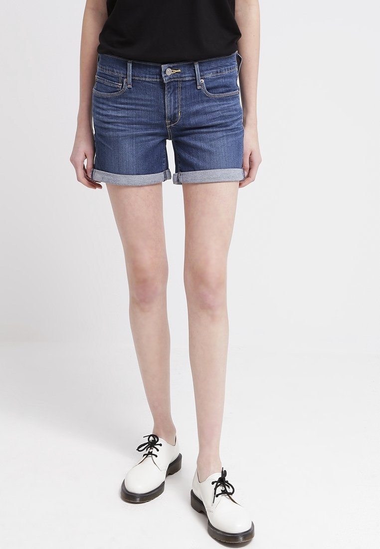 Person som bär uppkavlade blå jeansshorts, svart topp och vita snörskor, står mot en enkel ljus bakgrund.