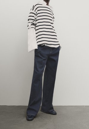 Personne portant une chemise blanche à manches longues sous un pull rayé noir et blanc, un jean large foncé et des mocassins noirs.