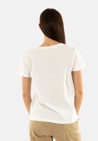 Camisa de algodón blanca con mangas cortas y escote redondo, que presenta un corte holgado y detalles de mangas enrolladas, combinada con pantalones beige.