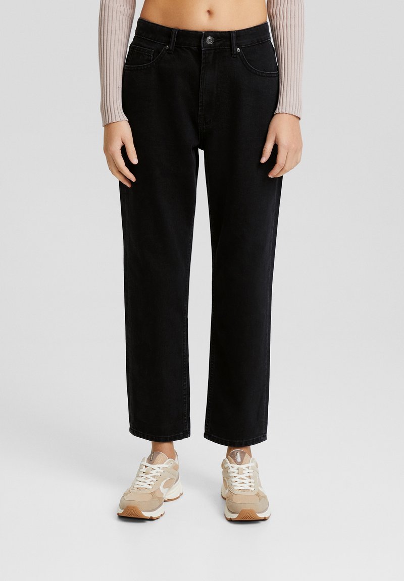 Personne portant un jean noir taille haute à jambes droites, des baskets beige et blanc, et un haut court beige côtelé à manches longues, debout sur un sol gris.