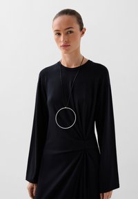 Abito lungo nero con maniche lunghe e un dettaglio di vita arricciata, abbinato a un grande pendente circolare argentato su un cordino nero. Tessuto dalla texture liscia.