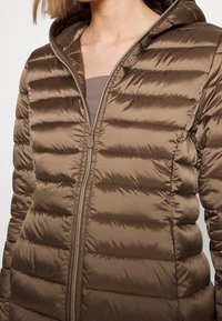 Braun glänzende gesteppte Jacke mit Kapuze, Reißverschluss teilweise geöffnet, über einem taupefarbenen Oberteil getragen, das den Ausschnitt und den oberen Brustbereich zeigt.
